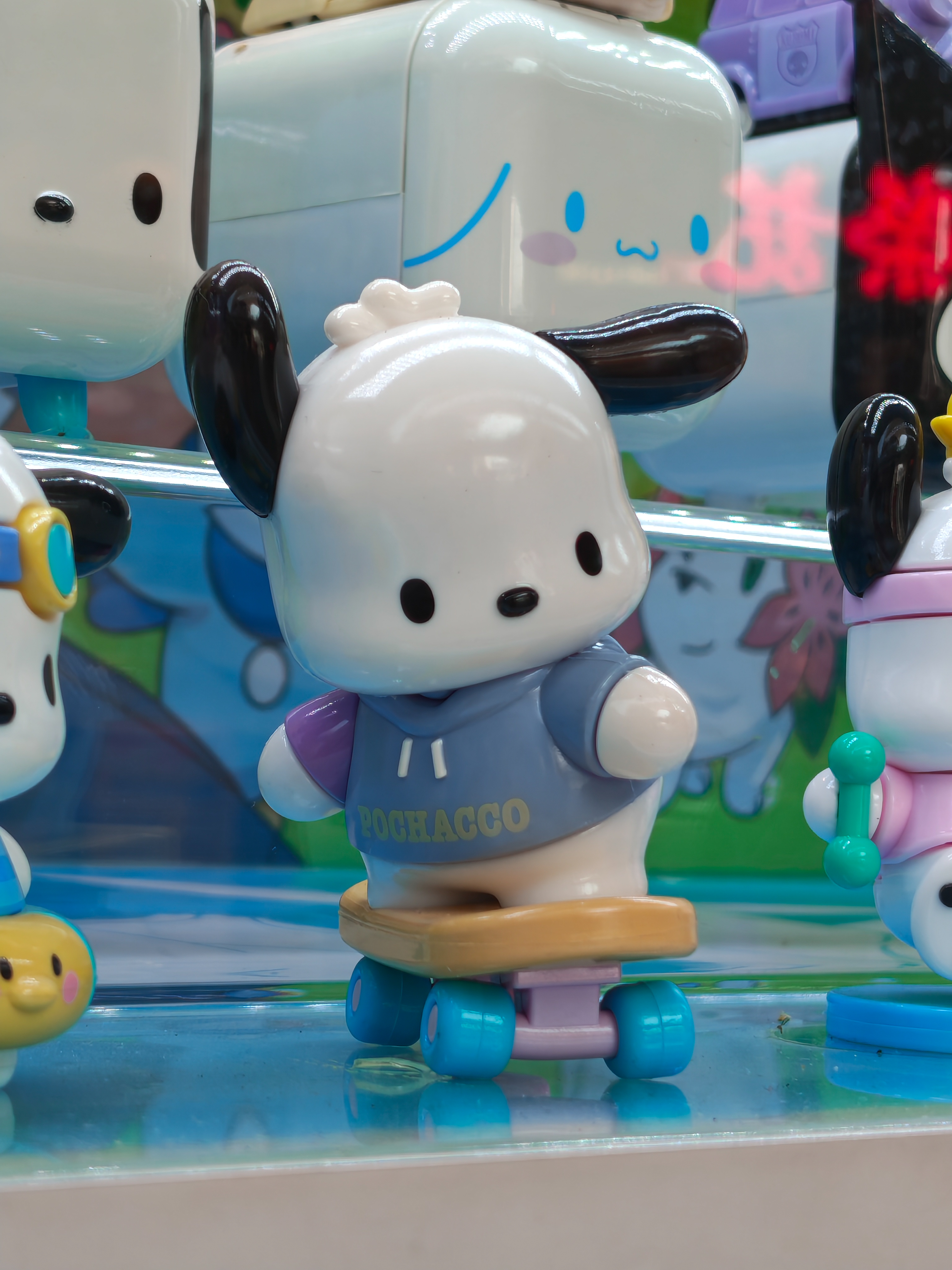 A macro photograph of a small plastic toy figure of the character Pochacco. The character is wearing a blue and purple hoodie that says "POCHACCO" and is standing on a small skateboard with blue wheels. The background is brightly colored and out of focus, suggesting a display case in a toy store.
