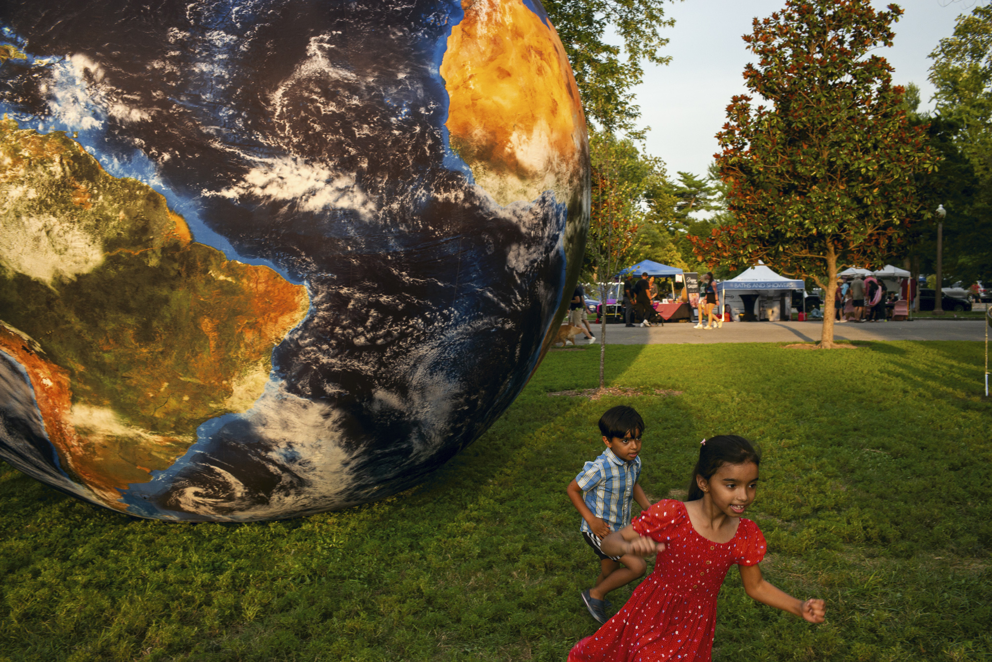 Two young children, a girl in a red dress and a boy in a checked shirt, run across a park lawn past a giant inflatable globe of the Earth, with market stalls and trees glowing in evening light