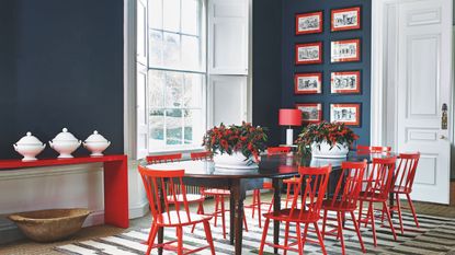 A blue and red dining room with a black table, red chairs, blue walls, and a black and red gallery wall. Beneath the table is a large striped rug, and to the left is a red console table with three white vases. Underneath is a wooden basket.
