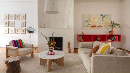 A white living room with a beige sofa, colorful pillows, a round coffee table, a beige armchair with a colorful striped throw, a side table, a wooden bunny, a floating orange sideboard and framed art