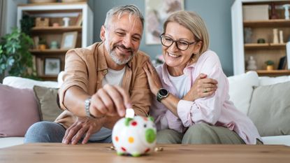Mature, smiling couple sitting on the couch and putting coins into a piggybank