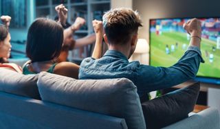 A group of football fans sitting on a sofa watching football on the TV