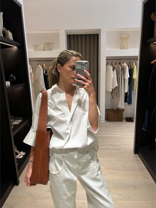 Eliza Huber in the dressing room at Banana Republic wearing a white poplin shirt and white baggy jeans with white flat slides and a brown leather oversize bag.