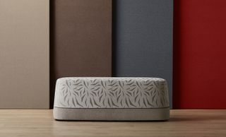 A gray and beige upholstered bench, with a darker gray pattern on it. Behind the bench are fabrics leaning on the wall.