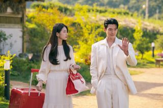 A woman (Bae Suzy) wears an all-white outfit and holds a red purse and a red suitcase, as a man (Kim Woo-bin) in all white stands next to her outdoors, in 'Genie, Make a Wish.'