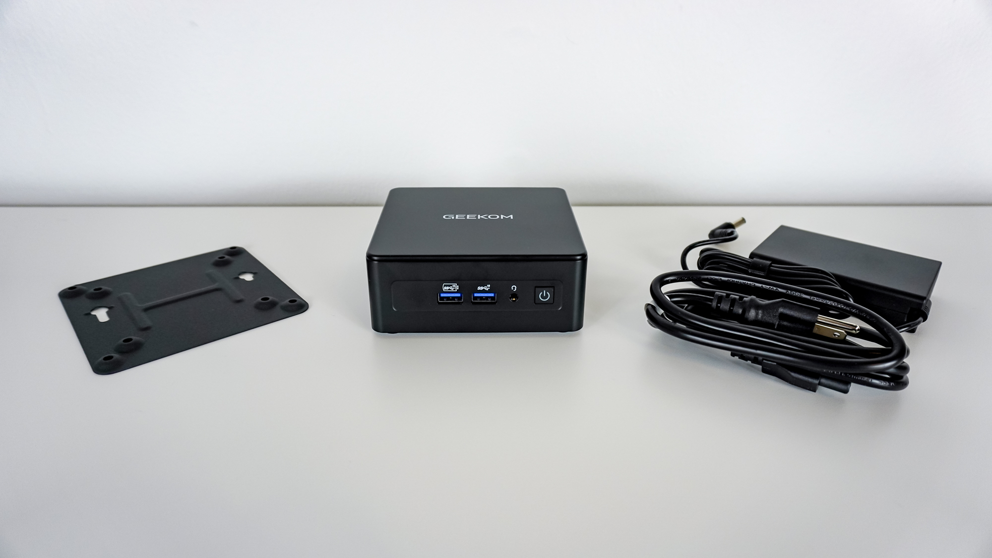The Geekom IT15 mini PC on a desk next to its power adapter and the included VESA mount plate adapter