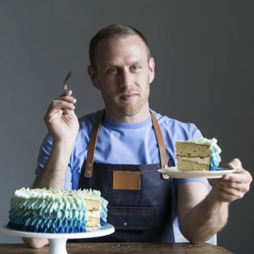 A headshot of a man in an apron eating a cake