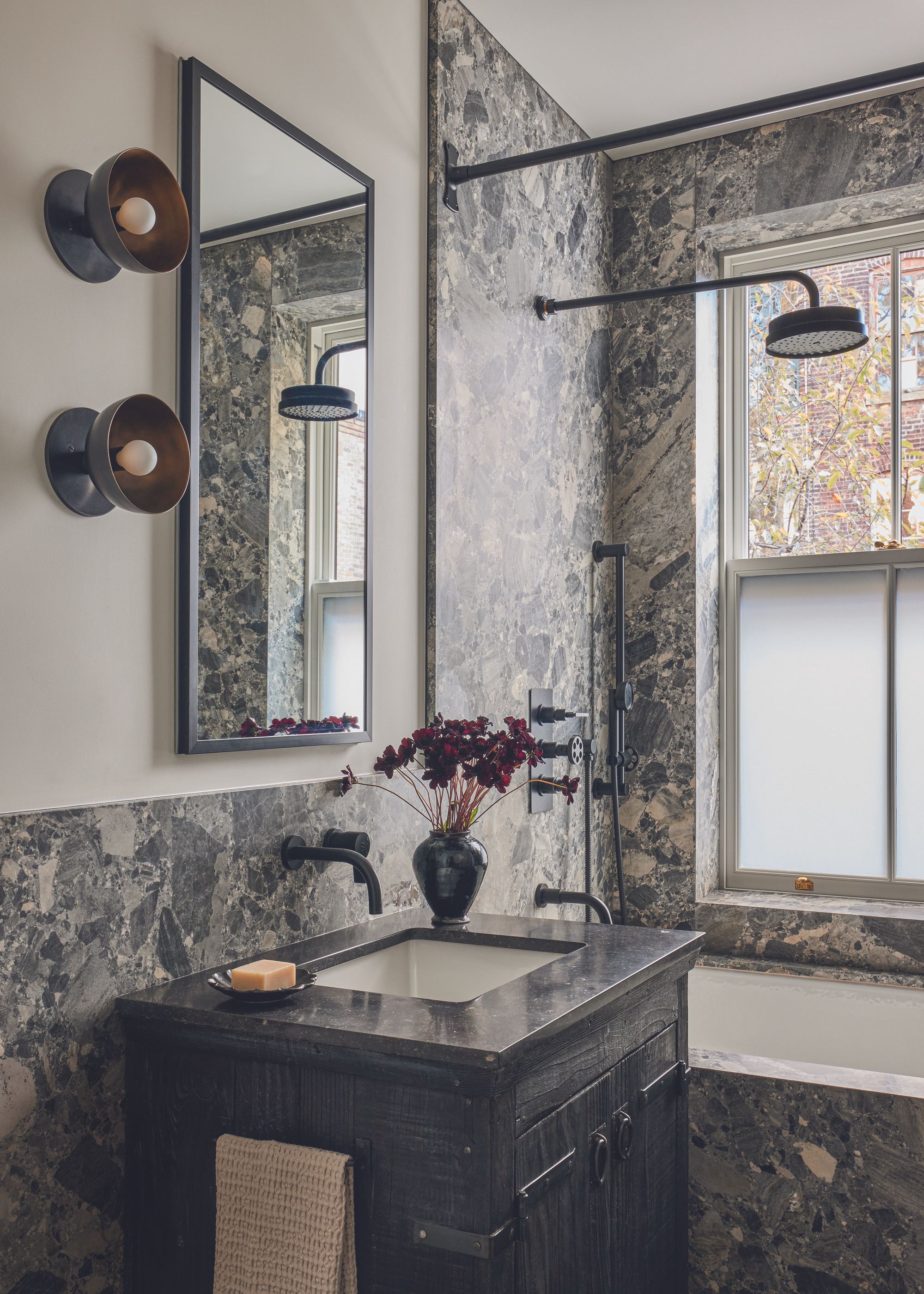 A dramatic dark stone bathroom featuring wall light fittings and stone covered walls and a small vase of flowers on the vanity unit