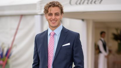 Alexander Ogilvy wearing a navy suit and pink tie and smiling in front of a white tent