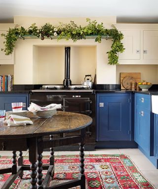 Kitchen with cream walls, bright blue units, black Aga with shelf overhead decorated with foliage and flowers, rounded dark wood dining table, red, blue and green floral rug beneath the dining table