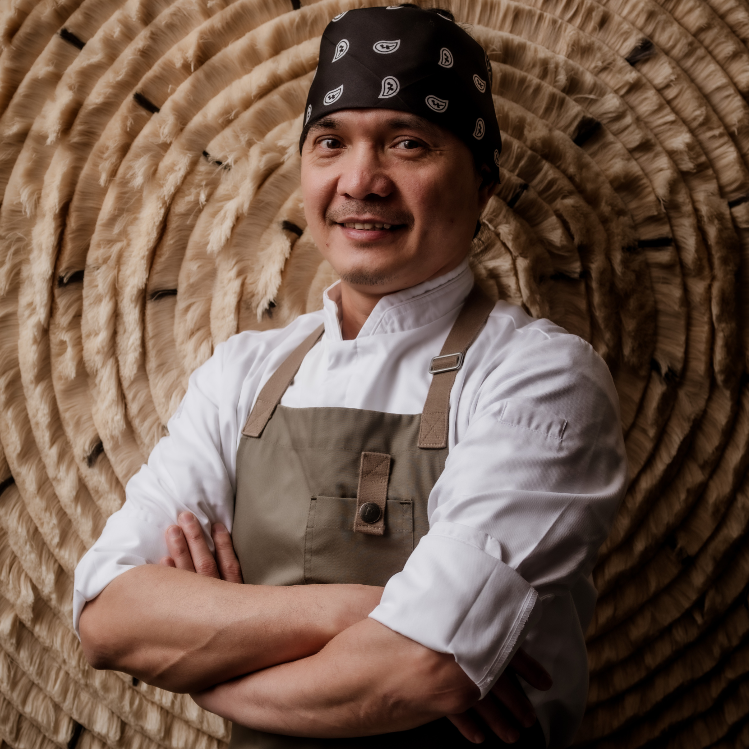 A headshot of a man in a paisley black bandana in a khaki green apron and white chef's jacket against a textured backdrop