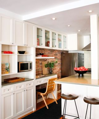A kitchen with white cabinets along the top wall and bottom shelves, a kitchen island with a wooden surface and dark brown wooden stools, a brick accent wall with a curved wooden chair underneath it