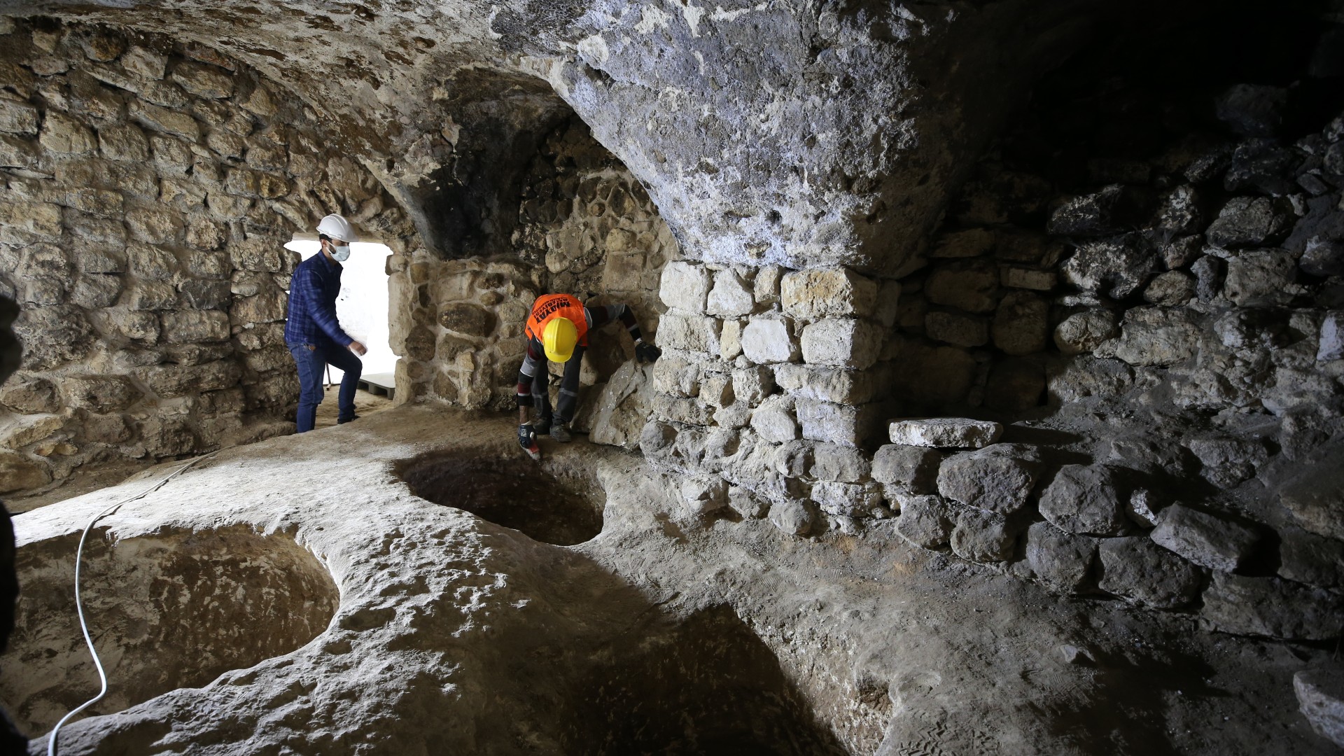 Two people who are wearing hard hats, masks and high visibility safety vests are exploring an underground cave thought to be a city. The walls are lined with large stone bricks and there are three circular holes on the floor.