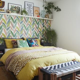 Bed with bedding, pillows and throws on it on wooden floor with leaf print wallpaper behind it