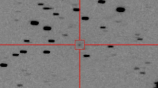 A black and white image shows a gray background speckled with dots with a crosshairs and a box circling a specific dot in the center of the image