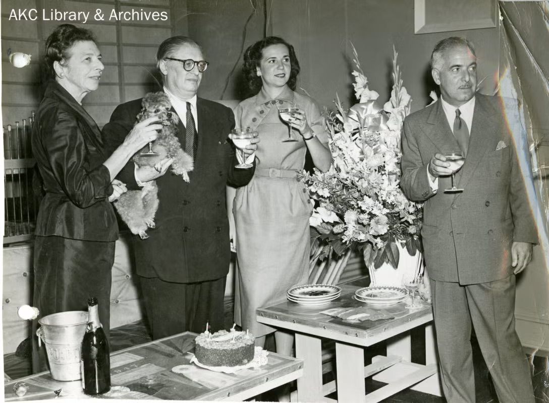 Count Alexis Pulaski stands with Mr and Mrs Gilbert Kahn and Nathalie Stuyvesant Pierrepont at a gathering, holding a toy poodle while guests raise glasses beside a table of flowers and champagne.