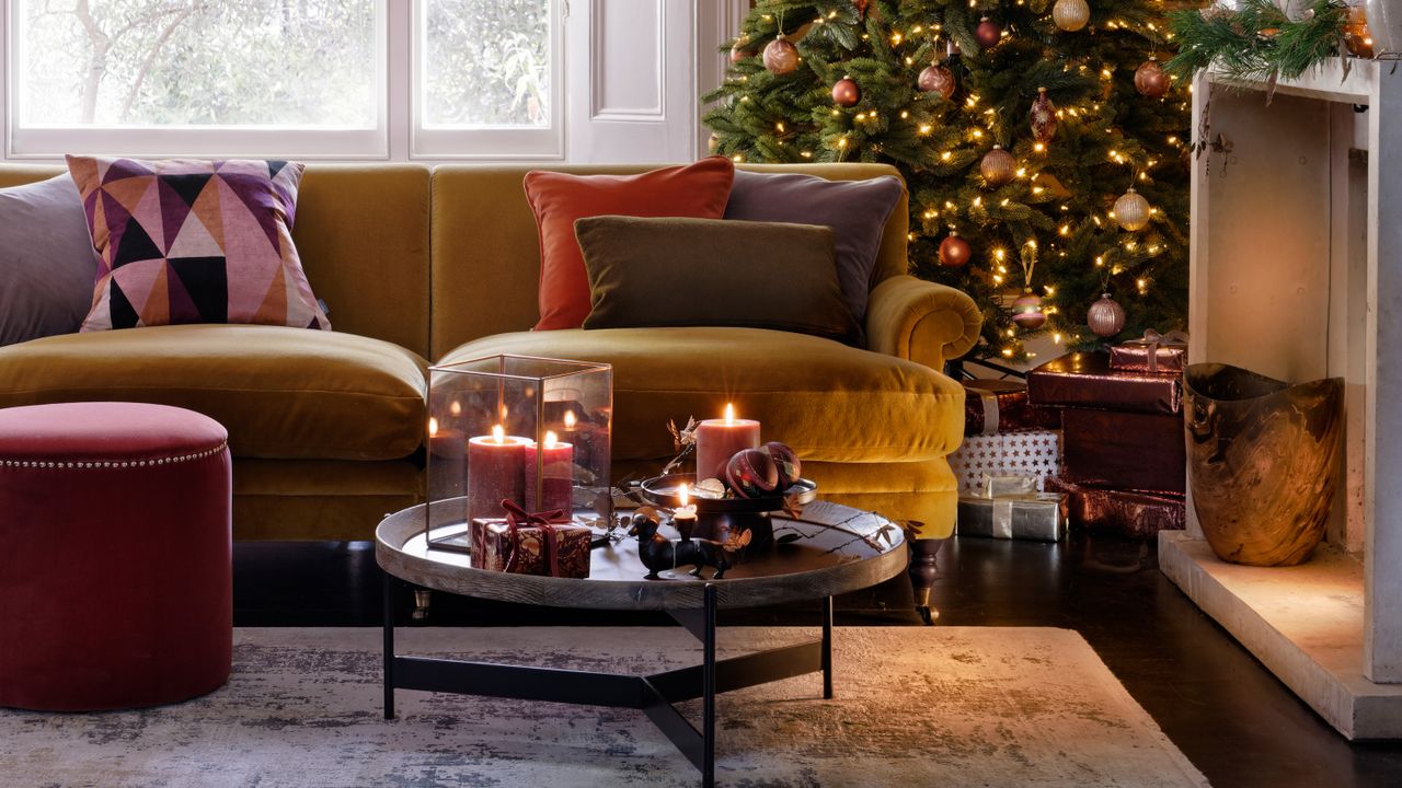 Gold velvet sofa lined with cushions behind a round coffee table with candles on top and a christmas tree with fairy lights in the background