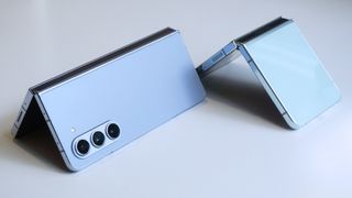 En ljusblå Samsung Galaxy Z Fold 5 ligger bredvid en Samsung Galaxy Z Flip 5 i samma färg, halvt öppnade på ett vitt bord.