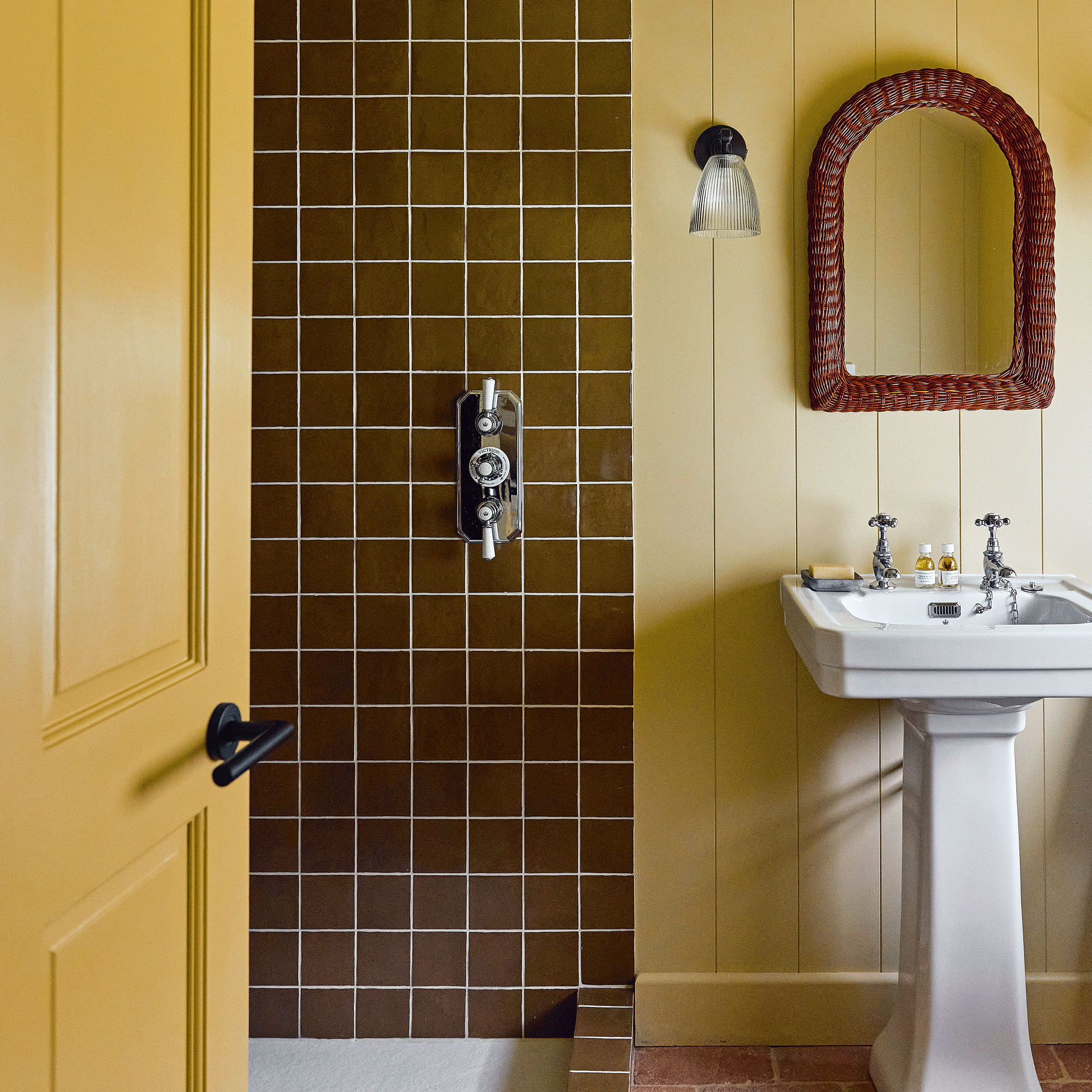 a yellow and brown bathroom with brown square tiles and yellow painted wall panelling, a traditional basin and wicker mirror