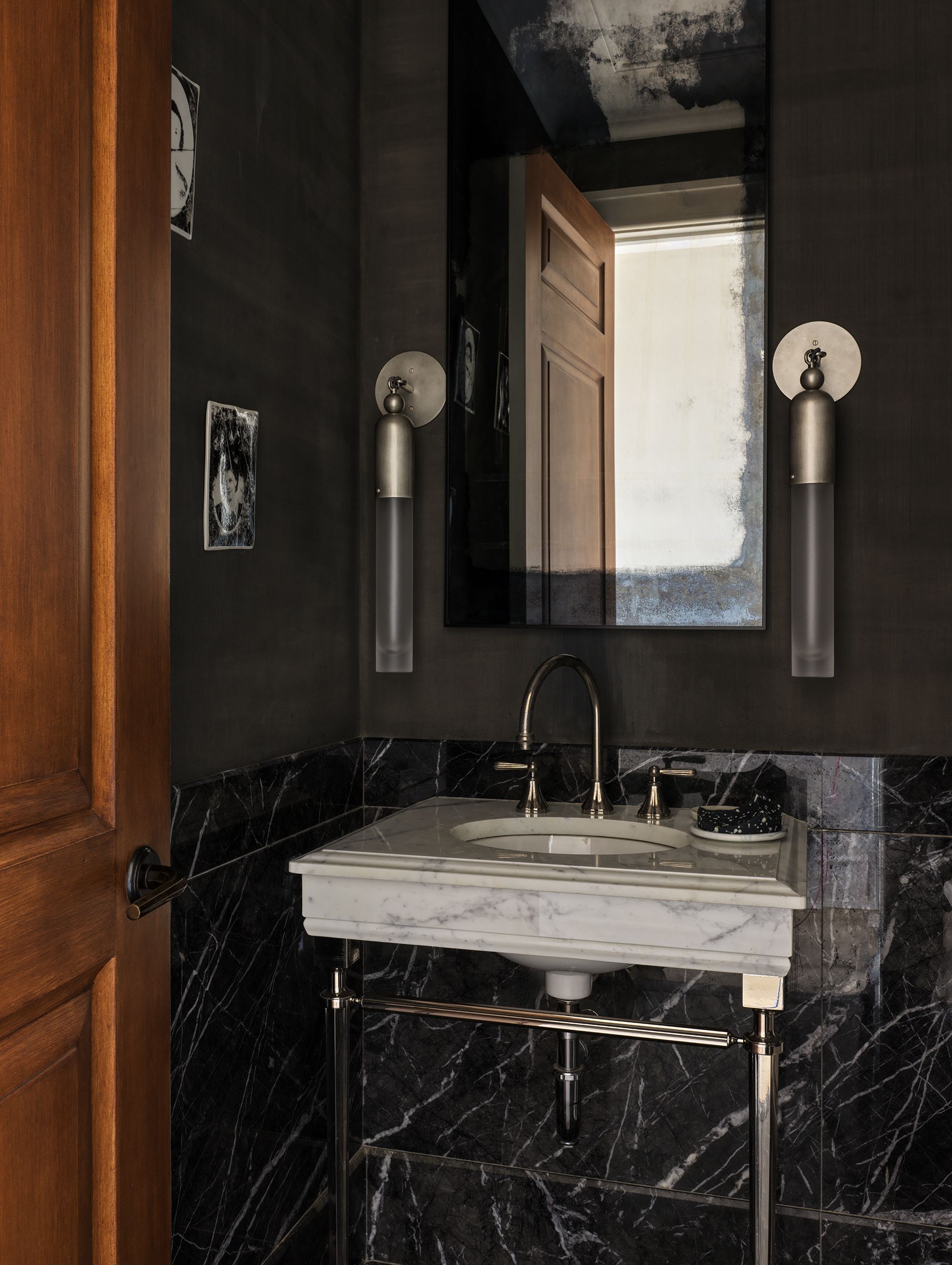 A powder room with dark walls and black marble and a white marble sink with slim brass legs below a mirror with a wall light either side of it