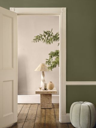 A green room with wooden floorboards with an open doorway leading to a taupe coloured hallway with a marble table and lamp
