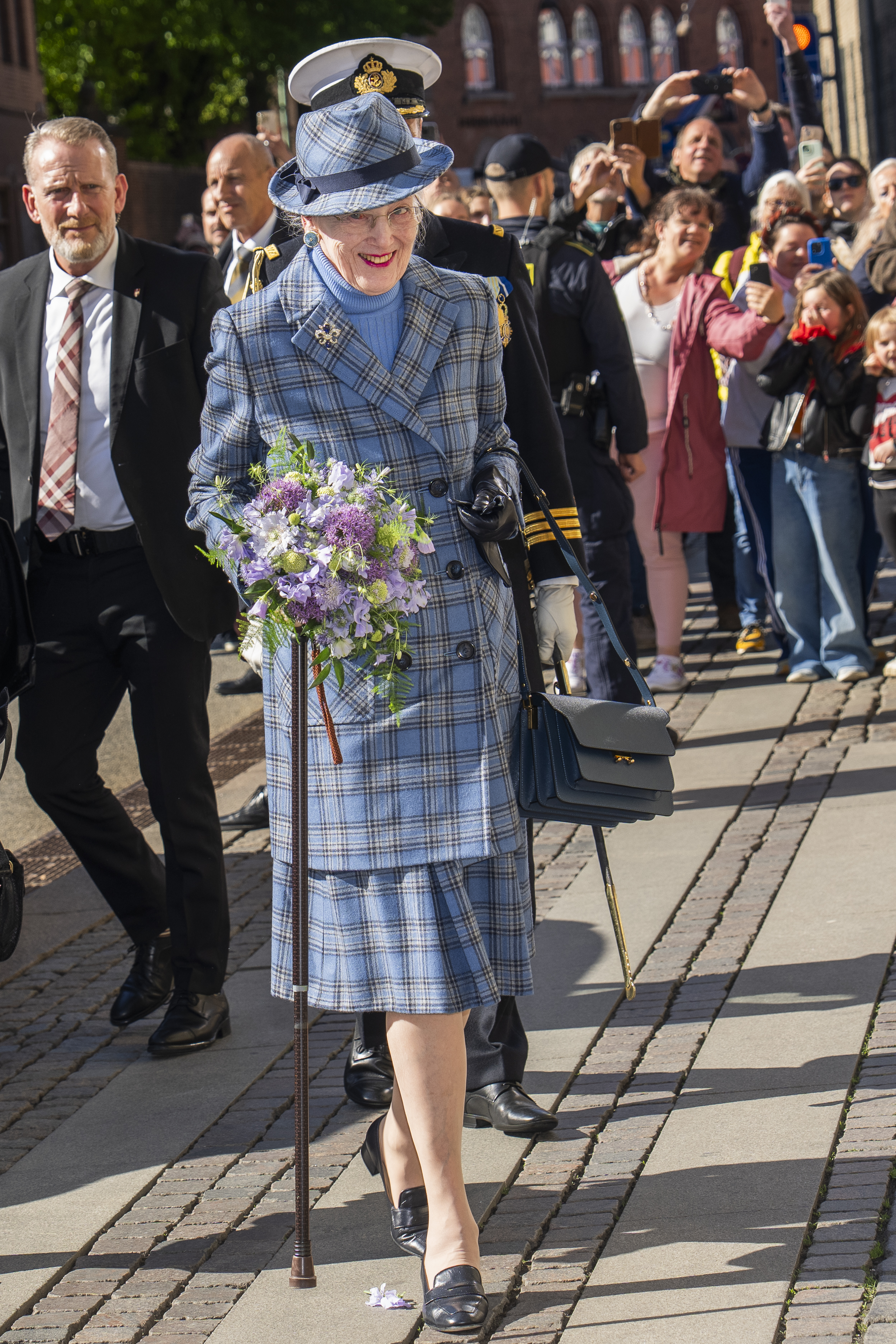 Queen Margrethe wearing a blue plaid suit and hat walking with a cane