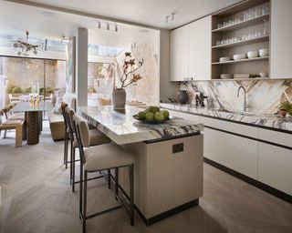 open plan kitchen with marble worktops, streamlined taupe cabinets, island with cream barstools and sitting and dining areas