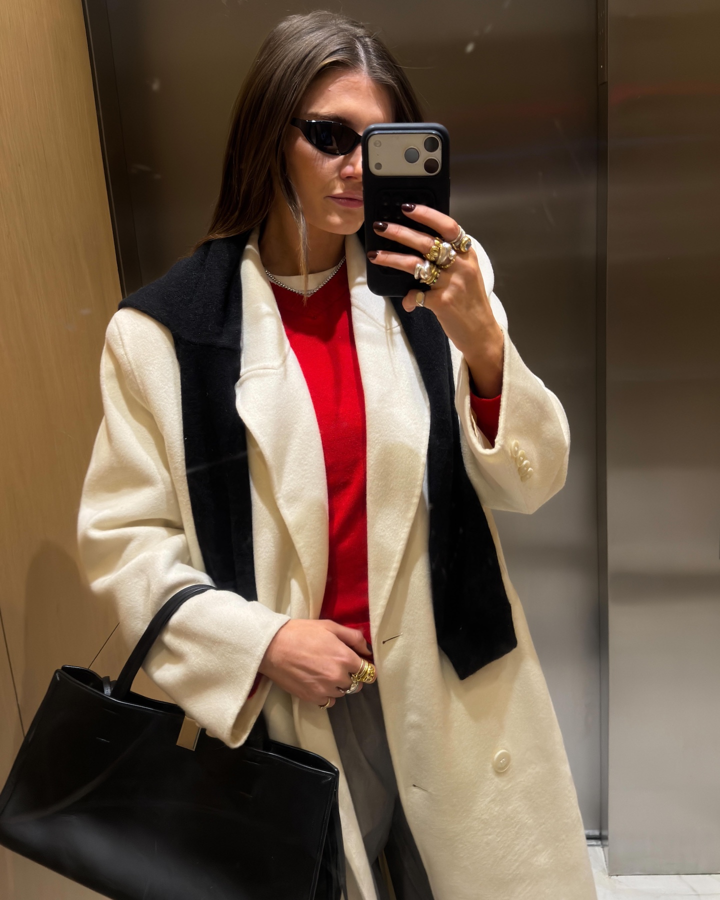 NYC style influencer Coco Schiffer poses for an elevator selfie wearing a chic outfit for 2026 with a cream coat and a red V-neck sweater layered over a white tee