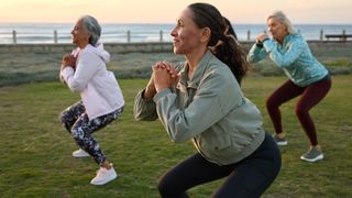Three mature women outdoors performing squats on the grass near the ocean at sunset