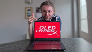 Ben Wilson of Windows Central points at a HyperX OMEN 15 laptop displaying a bright red &ldquo;OLED HYPERX&rdquo; screen while giving a thumbs‑up. The laptop has a multicolored backlit keyboard.