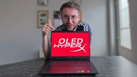 Ben Wilson of Windows Central points at a HyperX OMEN 15 laptop displaying a bright red &ldquo;OLED HYPERX&rdquo; screen while giving a thumbs‑up. The laptop has a multicolored backlit keyboard.