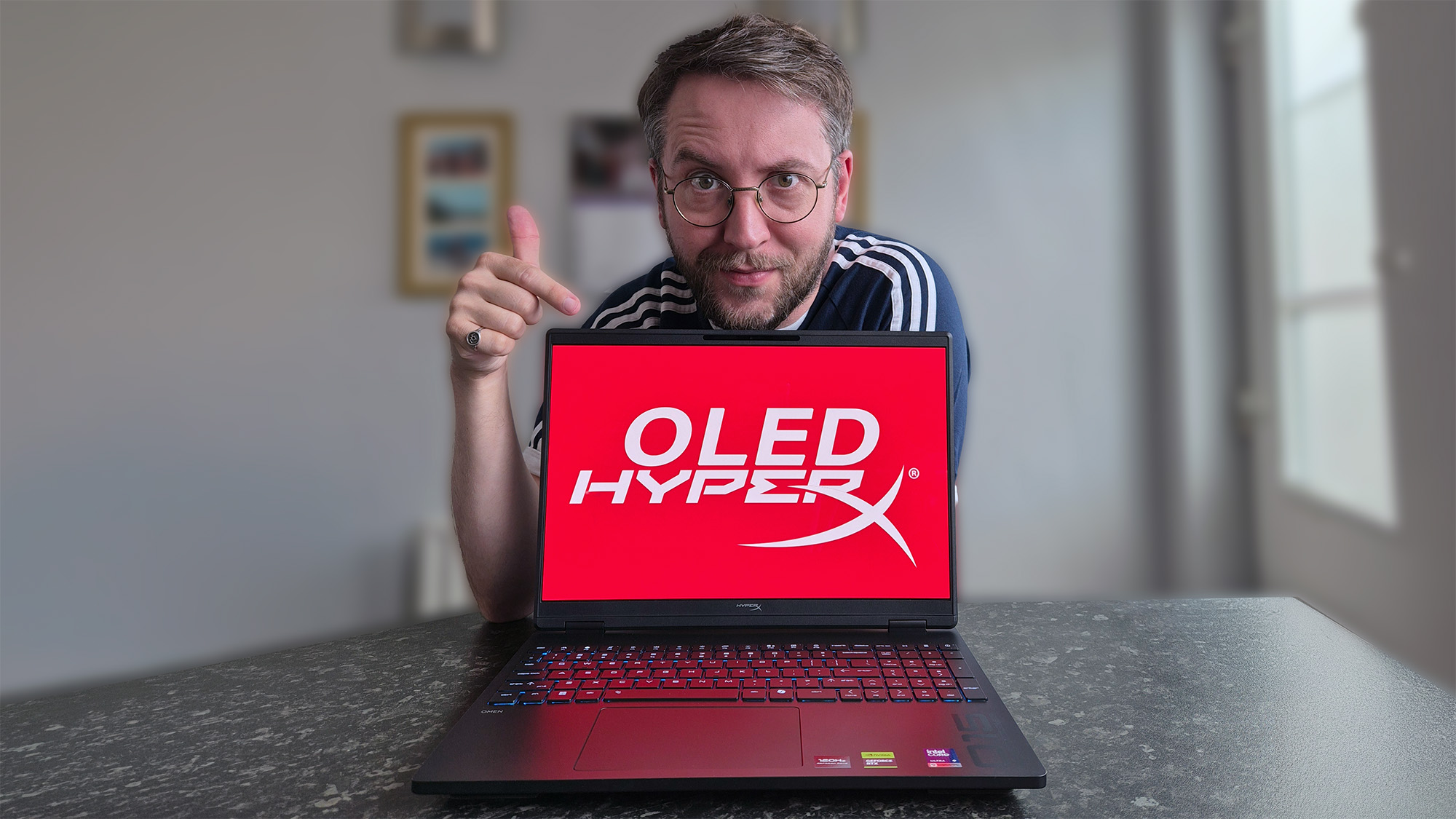 Ben Wilson of Windows Central points at a HyperX OMEN 15 laptop displaying a bright red &ldquo;OLED HYPERX&rdquo; screen while giving a thumbs‑up. The laptop has a multicolored backlit keyboard.