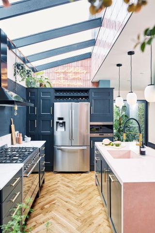 Kitchen with galley-style layout set in side return extension, with glass roof overhead, dark blue wall and units, and light pink island
