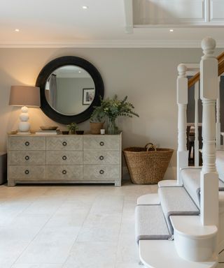 large hallway with cream stone flooring, dresser and large mirror above, wicker basket and white wooden staircase