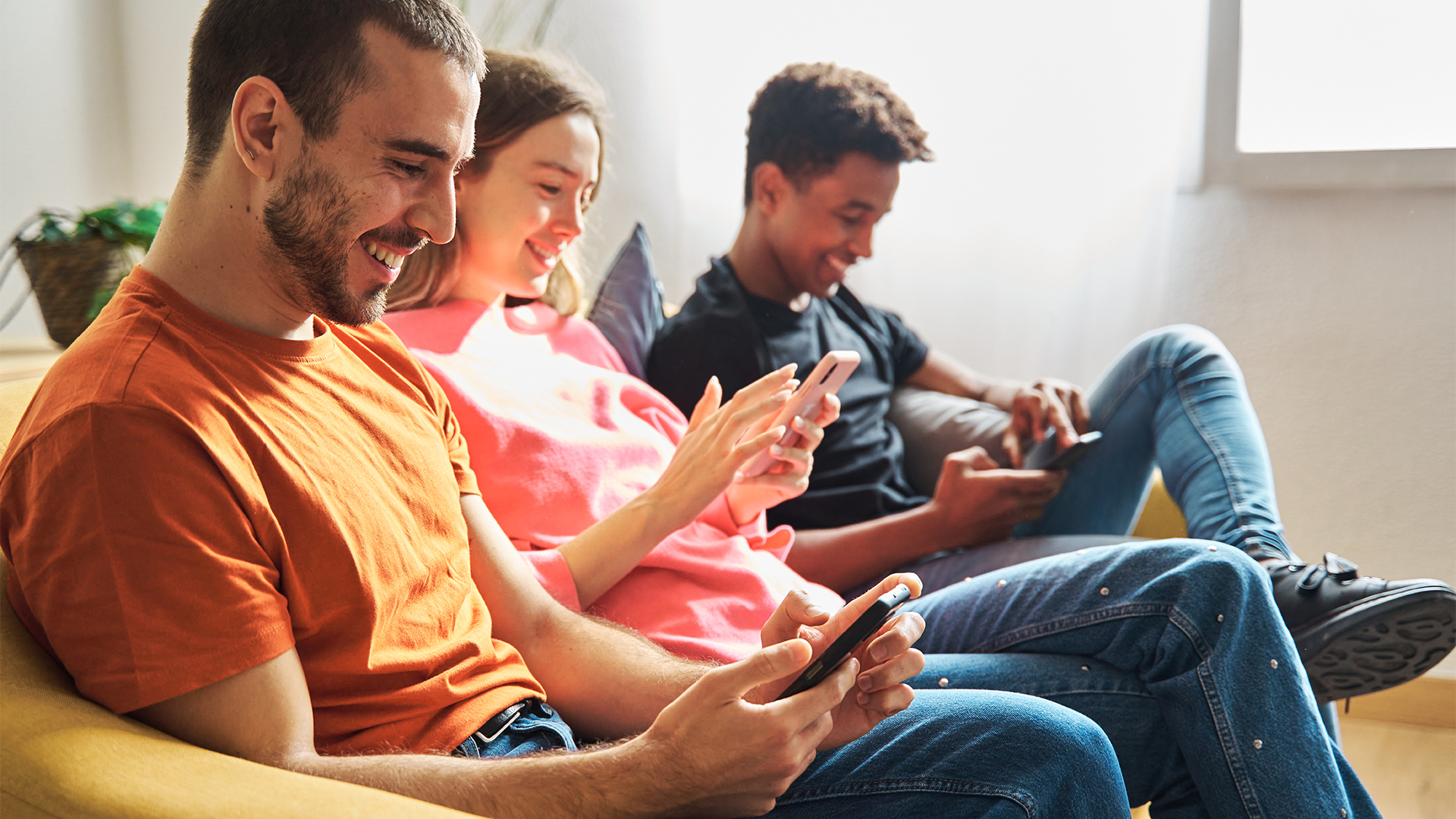 Multiracial group of roommates sitting in living room, smiling while checking social media on mobile phone.