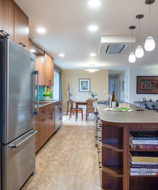 A narrow kitchen with brown cabinets, a silver fridge, a brown wooden kitchen island with books in it and with pendant lights over it, and a dining area in front of it