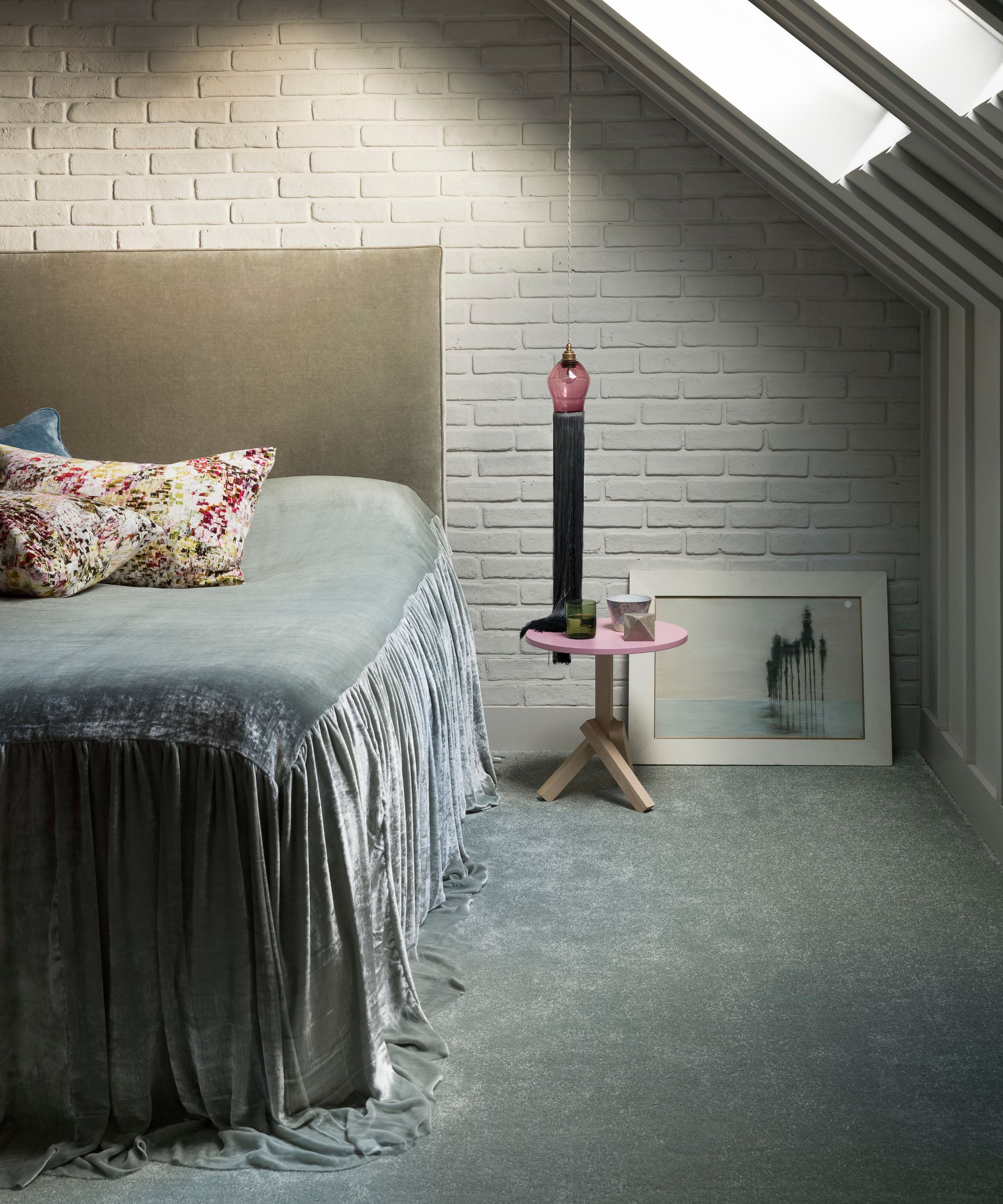 A minimalist attic bedroom with a white painted exposed brick wall, and large bed with velvet headboard, velvet throw, floral cushions and a pink bedside table, with a framed artwork in a white frame behind.