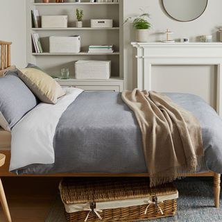 a bedroom with grey walls and inbuilt storage, with a bed that has a blue duvet cover with a brown cushion and blanket, with a wicket basket storage underneath the bed