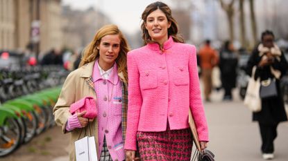 Two influencers in Paris, street styled outside of a fashion week show on a busy pavement. They are both wearing hot pink Chanel wool jackets with headbands and carrying Chanel beauty shopping bags