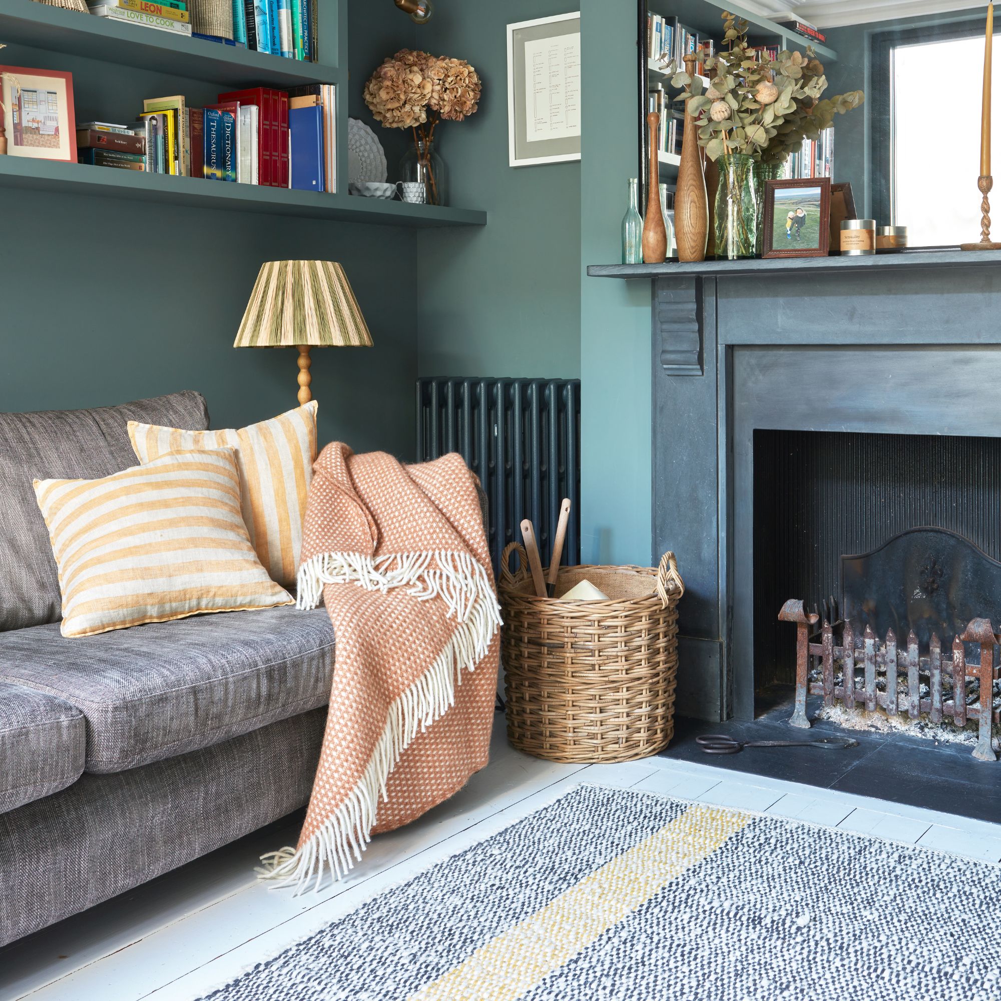 Teal painted living room with a period fireplace, and a grey sofa with cushions and a blanket on it