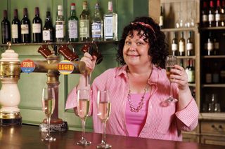 Gloria (Sue Vincent) stands behind the car at La Couronne, smiling at the camera with one hand on the pumps and the other holding up a glass of fizz