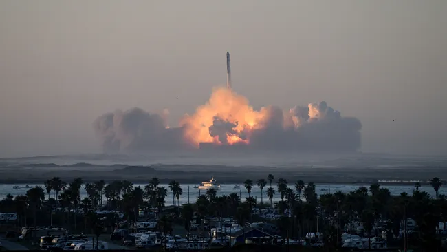 El lanzamiento de un cohete Starship de SpaceX desde la base de Boca Chica, Texas, al amanecer. El cohete se eleva hacia el cielo, dejando una estela de fuego y humo en la plataforma de lanzamiento. En primer plano, se pueden ver palmeras y una zona con vehículos y remolques, mientras que un barco navega en un cuerpo de agua cercano. La escena captura la magnitud del lanzamiento y el entorno costero desde donde fue realizado.