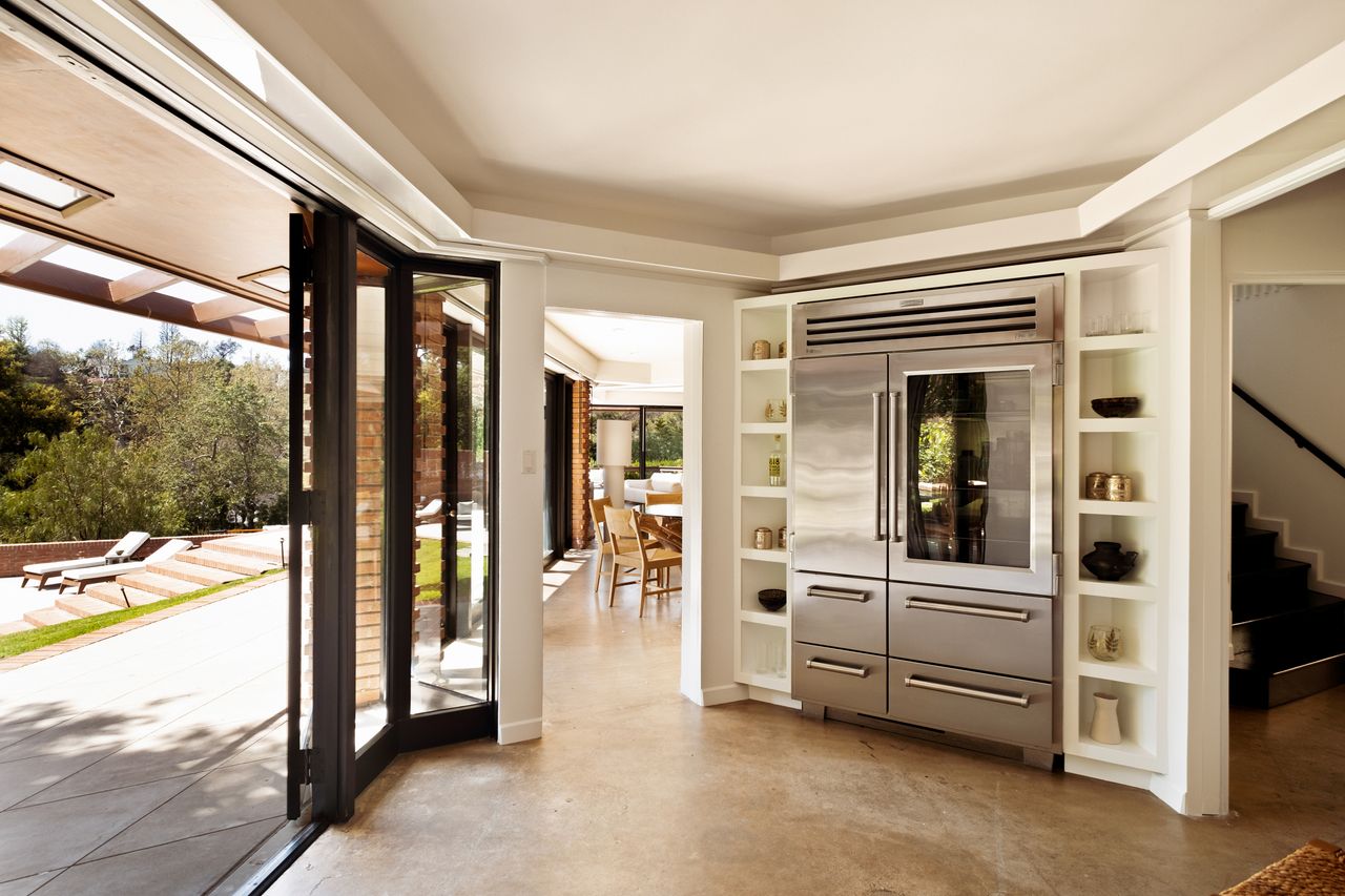 Image of an angularly-shaped kitchen with a large stainless steel fridge with built-in shelving on either side. There is a glass door open to the outside.