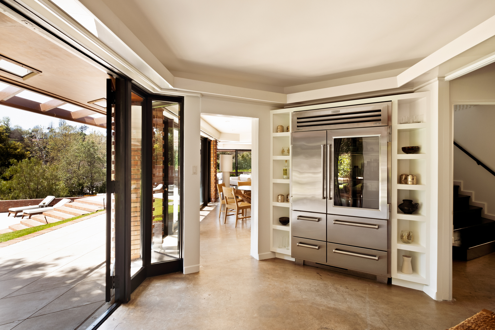 Image of an angularly-shaped kitchen with a large stainless steel fridge with built-in shelving on either side. There is a glass door open to the outside.