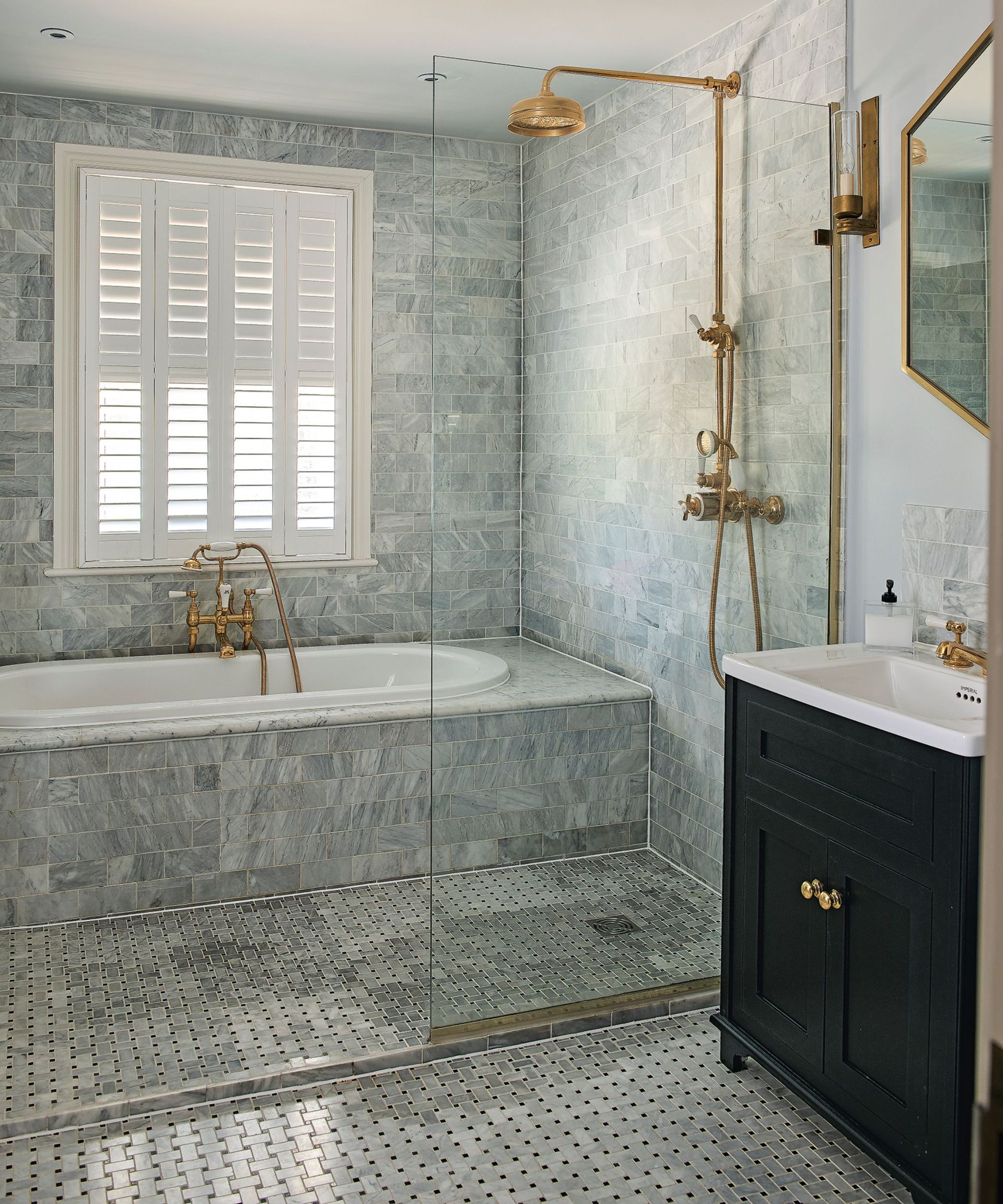 A tiled bathroom with a gold shower and gold faucets over a sunk tiled bath below a window with white shutters with a black cabinet with white porcelain countertop and gold hexagonal mirror