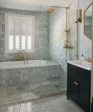 A tiled bathroom with a gold shower and gold faucets over a sunk tiled bath below a window with white shutters with a black cabinet with white porcelain countertop and gold hexagonal mirror