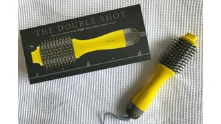 The Drybar Double Shot Blow Dryer Brush against a patterned white fabric background