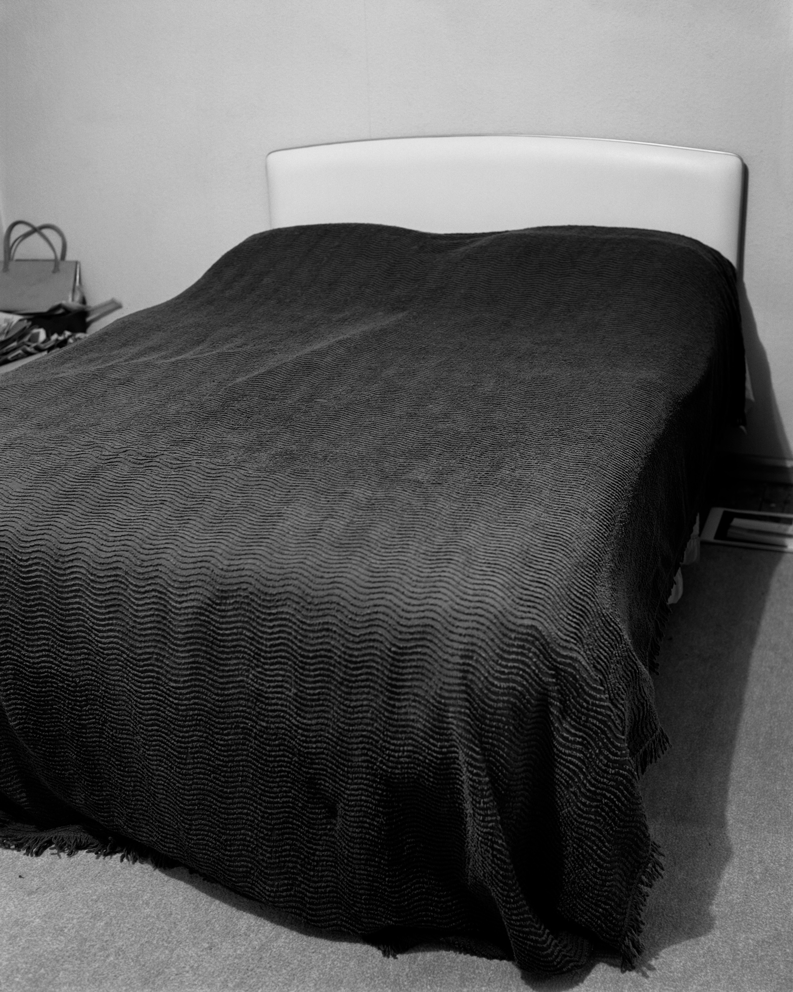 Black and white photograph of an unmade single bed with a dark, ridged bedspread pulled up against a plain headboard in a sparse room.