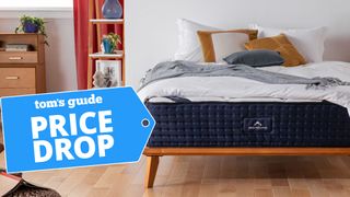 The DreamCloud Hybrid mattress placed on a wooden bedframe with a blue price drop sales badge overlaid on the image