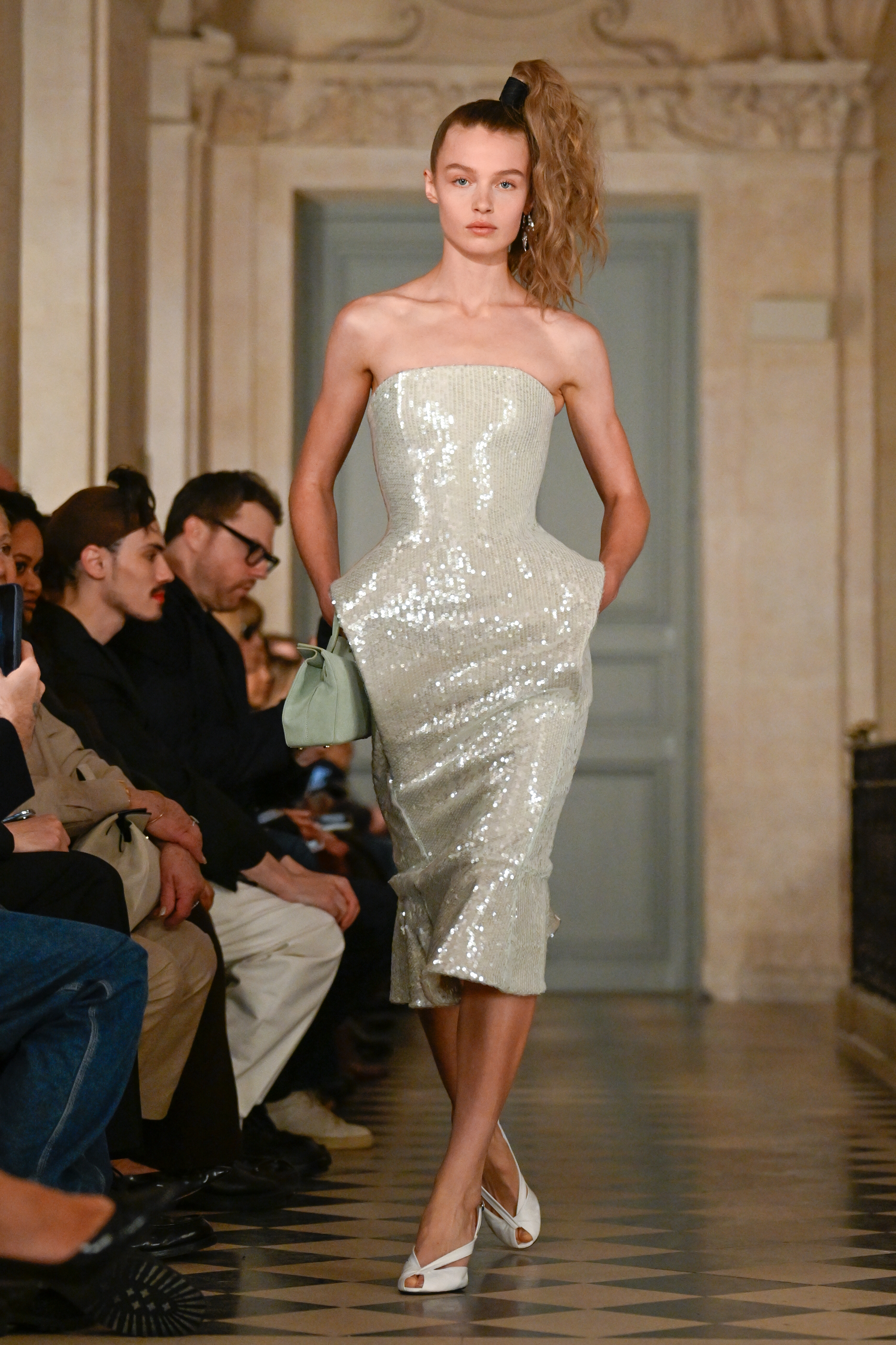 a model walks during a runway show wearing a strapless, sequin gown and white pumps. her hair is styled in a side ponytail